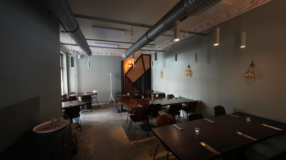Innenansicht eines modernen Restaurants mit langen dunklen Tischen und Stühlen in einem schmalen Raum. Sichtbare Lüftungsrohre an der Decke, hängende Pendelleuchten und graue Wände prägen das industrielle Ambiente.