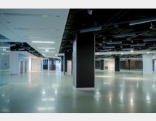 Leerer, moderner Innenraum eines Gebäudes mit glänzendem Boden, schwarzen Deckenpaneelen mit sichtbarer Verkabelung, weißen Wänden und Glaselementen. Mehrere Säulen und Notausgangsschilder sind sichtbar, die Umgebung wirkt offen und weitläufig.