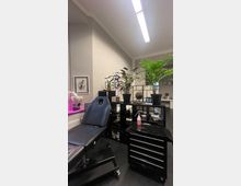 Innenraum eines modernen Studios mit einem schwarzen Behandlungsliege-Stuhl, einer Werkzeugkommode mit mehreren Schubladen und Pflanzen auf einem Regal. Die Wände sind beige gestrichen, und die Beleuchtung erfolgt durch eine helle Deckenleuchte.