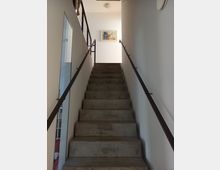 Eine gerade Treppe aus grauem Beton mit schwarzen Metallgeländern führt zu einem hell beleuchteten Flur. An der weißen Wand oben hängt ein buntes Gemälde, und links ist ein Wandspiegel zu sehen.