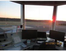 Blick aus einem Kontrollturm eines Flughafens mit großen Fenstern, die die Start- und Landebahn sowie umliegende Felder und Wälder bei Sonnenuntergang zeigen. Im Vordergrund sind mehrere Monitore, Schreibtischutensilien und Kommunikationsgeräte sichtbar.