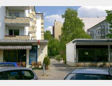 Mehrstöckiges Wohngebäude mit Balkonen aus gelblichen Ziegeln links, daneben ein kleiner Laden mit gestreifter Markise und Sitzgelegenheiten auf dem Gehweg. Im Vordergrund parken Autos, und im Hintergrund sind grüne Bäume sowie ein schmaler Weg zwischen Gebäuden zu sehen.