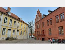 Straßenszene mit historischen Gebäuden: Links ein hellgelbes Haus mit weißer Tür und Fenstern, rechts ein rotes Backsteingebäude im gotischen Stil mit spitzbogigen Fenstern. Dazwischen ein gepflasterter Platz mit einigen Menschen und Fahrrädern, im Hintergrund Bäume und kleinere Gebäude.