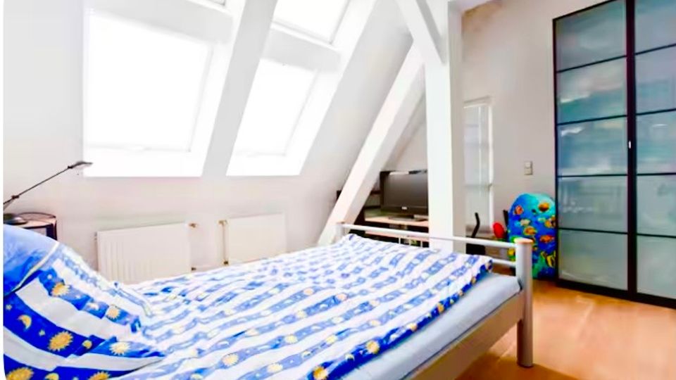 Helles Schlafzimmer im Dachgeschoss mit schrägen Decken, großen Dachfenstern und weißen Sichtbalken. Im Raum stehen ein Bett mit blau gemusterter Bettwäsche, ein großer Kleiderschrank mit Schiebetüren sowie ein kleiner TV-Bereich auf hellem Holzboden.