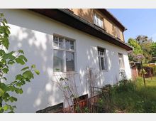 Zweistöckiges Wohnhaus mit weißer Fassade im unteren Bereich und braunem Putz im oberen Bereich, umgeben von verwildertem Garten und Vegetation. Es sind mehrere Fenster zu sehen, und im Vordergrund wachsen Sträucher sowie hohe Gräser.