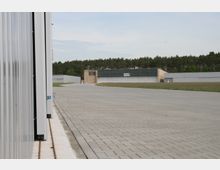 Gepflasterte Fläche mit angrenzenden Industriegebäuden, darunter ein großer Hangar und weitere Hallen, im Hintergrund von einer Baumreihe begrenzt. Links im Bild ist eine metallene Gebäudewand mit einer Schienenführung erkennbar.