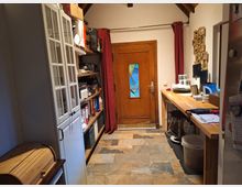 Ein schmaler Raum mit einer Holztür, die ein buntes Glaselement hat, und roten Vorhängen an den Seiten. Links steht ein weißer Glasschrank und ein Regal mit Büromaterialien, rechts befindet sich eine lange Holzarbeitsplatte mit Barhockern und Haushaltsgeräten. Der Boden besteht aus Fliesen im Naturstein-Look.