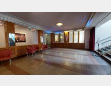 Empfangsbereich eines Gebäudes mit hölzernem Tresen und Wandverkleidung. Es gibt rote Stühle und kleine Tische, eine beleuchtete Treppe auf der linken Seite und einen Zugang zu weiteren Räumen auf der rechten Seite.