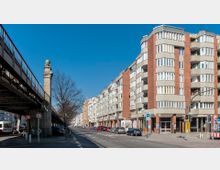 Straßenansicht in einer Stadt mit mehrstöckigen Wohnhäusern aus Backstein und Beton, angeordnet entlang einer breiten Straße. Links im Bild verläuft eine erhöhte Brücke mit Geländer; Autos parken entlang der Straße.