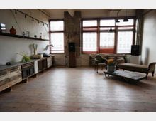 Ein industrial-inspirierter Raum mit Holzboden, großen Fenstern mit Holzjalousien und einer Kombination aus alten und rustikalen Möbeln, darunter ein Sideboard aus recyceltem Holz, ein Sofa, ein Sessel und ein Couchtisch auf Rollen. Im Hintergrund sind freiliegende Ziegelwände und hängende Deckenlampen zu sehen.