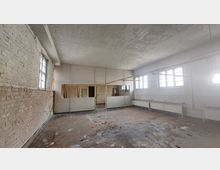 Leerer Raum mit abgenutztem Boden, unverputzten Ziegelwänden und einer Decke mit abblätterndem Putz. Große Fenster lassen Tageslicht herein, und die Wand gegenüber ist mit Trennwänden und mehreren Türen ausgestattet.