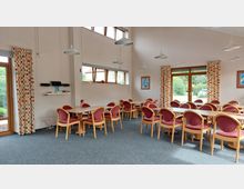 Ein heller Gemeinschaftsraum mit mehreren Holztischen und gepolsterten Stühlen mit rotem Stoffbezug. Große Fenster und Glastüren lassen viel Tageslicht herein, ergänzt durch farbenfrohe Vorhänge mit Karomuster; an den Wänden hängen Bilder und eine kleine Regalfläche mit elektronischen Geräten.