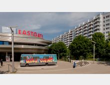 Außenansicht eines Einkaufszentrums mit großem roten Schriftzug „EASTGATE“ auf einer modernen, geschwungenen Metallfassade. Im Vordergrund ein bunt gestalteter Bus mit Graffiti sowie eine gepflasterte Fläche; dahinter Wohngebäude und grüne Bäume.