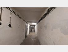 Langer, schmaler Korridor mit abblätternden weißen Wänden und mehreren schweren, metallverkleideten Türen auf der linken Seite. An beiden Wänden verlaufen sichtbare Rohre und Kabel, die Beleuchtung erfolgt über einfache, hängende Deckenlampen.