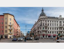 Straßenszene in einer Stadt mit einem historischen, weißen Eckgebäude mit reich verzierter Fassade und Kuppel auf der rechten Seite, sowie einem schlichten, mehrstöckigen Gebäude in Gelb und Rosa auf der linken Seite. Im Vordergrund befindet sich eine belebte Kreuzung mit Autos und Fußgängern, sowie einem U-Bahn-Schild für die Linie U7.