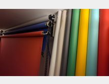 Mehrere farbige Hintergrundrollen, darunter Rot, Blau, Weiß, Grün, Gelb und Grau, sind an einer Wandhalterung montiert. Die Rollen sind teilweise ausgerollt und miteinander verbunden, typischerweise verwendet in Fotostudios.