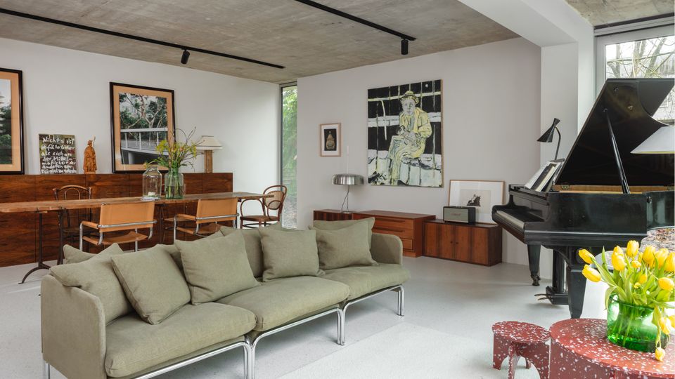 Modern open-plan living and dining room with exposed concrete ceiling, white walls, and large windows, featuring a green modular sofa on a light rug and a black grand piano. A long wooden dining table with chairs, low wood cabinets, and framed artwork line the walls, with a small red side table holding a vase of yellow flowers.
