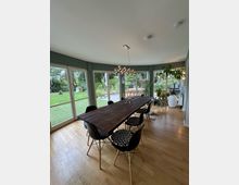 Wohnraum mit großem Esstisch aus dunklem Holz und schwarzen Stühlen, umgeben von großen Fenstern, die einen Blick auf einen Garten bieten. Der Raum hat einen Holzboden, grüne Wände und wird von einer modernen Deckenlampe sowie verschiedenen Zimmerpflanzen dekoriert.