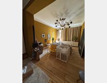 Wohnzimmer mit senfgelben Wänden und Holzboden, eingerichtet mit einer Couch, einem Babybett, einem gelben Beistelltisch und einem hängenden Korbsessel. Im Raum befinden sich ein moderner Kronleuchter, kleine Pflanzen und bodenlange beigefarbene Vorhänge vor einem Fenster.