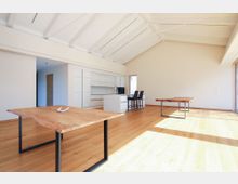 Heller, moderner Wohnraum mit hohen, weißen Deckenbalken und großen Fenstern, die Tageslicht auf den hölzernen Boden werfen. Der Raum enthält eine offene Küche mit weißen Schränken, zwei minimalistische Holztische und einen Essbereich mit Hochstühlen.