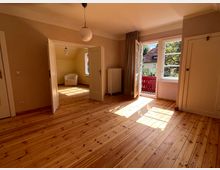 Helles Zimmer mit Holzdielenboden und großen Fenstern, die viel Tageslicht hereinlassen. Eine Doppeltür führt in einen angrenzenden Raum, und eine weitere Tür öffnet sich zu einem Balkon mit rotem Geländer und Blick auf Bäume und Nachbarhäuser.