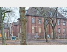 Backsteingebäude mit mehreren Fensterreihen, teils mit verschlossenen Rollläden und Brettern, umgeben von einem Bauzaun und einzelnen Bäumen im Vordergrund auf einem bewaldeten Gelände. Im Hintergrund sind weitere Gebäude im ähnlichen Stil zu sehen.