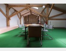 Ein Sitzungsraum mit grüner Teppichfläche, einer zentralen Holztafel und umstehenden Stühlen. Die Raumdecke wird von sichtbaren Holzbalken gestützt, und eine Ziegelwand ist im Hintergrund zu sehen.