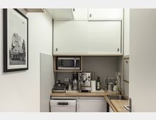 Kleine Küchenzeile mit weißem Schrank, eingebauter Mikrowelle und Kaffeevollautomat auf der Arbeitsfläche. An der Wand hängt ein gerahmtes Schwarz-Weiß-Bild; Spüle und Küchengeräte wie Toaster und Wasserkocher sind ebenfalls sichtbar.