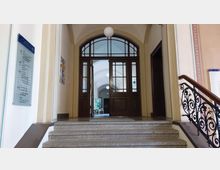 Treppenaufgang in einem historischen Gebäude mit verzierten Geländern aus Metall, einer Glastür mit Holzrahmen und einem dahinterliegenden Flur mit Rundbögen und Hängelampen. Rechts hängt eine Übersichtstafel mit Etageninformationen.