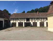 Ein Innenhof mit einem Fachwerkgebäude, das ein ziegelgedecktes Dach und mehrere Holztüren aufweist. Vor dem Gebäude befindet sich ein gepflasterter Boden, und im Hintergrund stehen Bäume unter klarem, blauem Himmel.