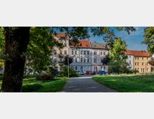 Ein Park mit einem Weg, der von Grasflächen und großen Bäumen gesäumt ist, im Vordergrund; im Hintergrund stehen historische Wohnhäuser mit verzierten Fassaden und roten Dächern. Autos sind vor den Gebäuden geparkt.