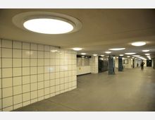 Ein unterirdischer Durchgang mit weißen, gefliesten Wänden und blauer Fliesenverkleidung an den Säulen. Die Decke ist mit runden, leuchtenden Deckenlampen ausgestattet, und im Hintergrund sind geschlossene Metalltüren sowie zwei Personen sichtbar.