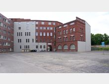 Backstein- und Putzgebäude in einem rechteckigen Innenhof mit großen Fenstern und einem Torbogen in der Mitte; rechts ein kleiner Parkplatz mit einem blauen Parkschild und im Hintergrund grüne Bäume.