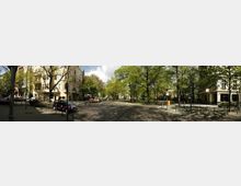 Straßenszene in einer Wohngegend mit Altbauten, umgeben von vielen Bäumen und einem grünen Platz in der Mitte. Parkende Autos und Gehwege mit Pollern säumen die gepflasterte Straße.