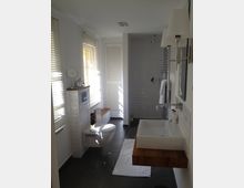 Modernes Badezimmer mit weißen Wandfliesen, dunklem Boden und Tageslicht durch Fenstervorhänge. Zu sehen sind ein wandhängendes WC, ein Waschbecken mit Holzablage, eine bodenebene Dusche und ein Handtuchhalter.