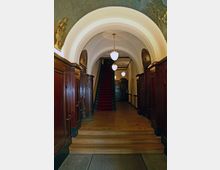 Eingangsbereich eines historischen Gebäudes mit Holzvertäfelungen, einer Treppe mit rotem Teppich und Rundbögen. An der Decke befinden sich Gemälde und hängende Kugelleuchten, die den Raum beleuchten.