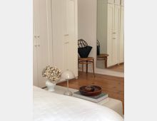 Helles Schlafzimmer mit weißen Einbauschränken, einem Bett im Vordergrund und einem Holzboden. Auf einem Hocker steht eine schwarze Tasche, daneben ein großer Spiegel; ein Beistelltisch zeigt Blumen, eine Lampe und Dekoration.