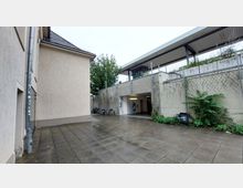 Gepflasterter Hof mit Zugang zu einem Unterführungstunnel, umgeben von einem Gebäude mit beigem Putz und einer Betonwand, die mit Pflanzen und Klettergewächsen bewachsen ist. Im Hintergrund ist ein überdachter Bereich mit Metallgeländern und Grünflächen sichtbar.