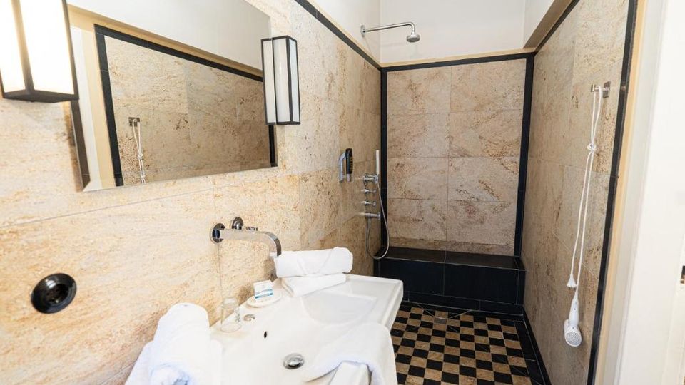 Modernes Badezimmer mit beigen Natursteinfliesen und schwarzer Rahmenkante, großem Wandspiegel und Waschbecken im Vordergrund. Hinten befindet sich eine Dusche mit Wand- und Handbrause sowie ein schwarz-weiß kariert gefliester Boden.