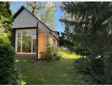 Einfamilienhaus mit Holz- und Metallfassade, großen bodentiefen Fenstern und einem steil geneigten Dach, umgeben von einem Garten mit Rasen, Bäumen und Sträuchern. Im Hintergrund ist eine Terrasse mit Gartenmöbeln sichtbar.