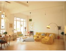 Helles Wohnzimmer mit großen Fenstern, gelber Polstermöbelgruppe und modernen Einrichtungsgegenständen. Im Raum befinden sich eine Stehlampe, ein Teppich, ein Esstisch mit Stühlen und Pflanzen als Dekoration.