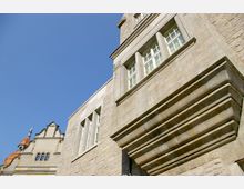 Historisches Gebäude mit Sandsteinfassade und großen rechteckigen Fenstern, daneben ein Teil eines zweiten Gebäudes mit dekorativem Giebel und orangem Spitzdach; blauer Himmel im Hintergrund.