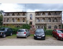 Ein alter, dreistöckiger Fachwerkbau mit Verzierungen und teilweise verfallener Fassade; vor dem Gebäude sind mehrere geparkte Autos auf einem unbefestigten Parkplatz zu sehen, umgeben von Büschen und Grünpflanzen.