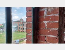 Blick durch eine Ziegelsteinmauer auf ein historisches Backsteingebäude mit symmetrischen Fenstern, verziert mit hellgrünen Elementen. Im Vordergrund eine grüne Wiese mit einer Steinmauer und einem Strandkorb, unter einem teils bewölkten Himmel.