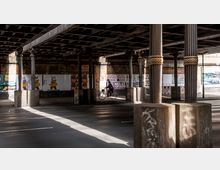 Unterführung mit verzierenden Metallsäulen, die ein Gitterdach stützen. Im Hintergrund ist eine Backsteinmauer mit Plakaten bedeckt, während eine Person auf einem Fahrrad durch den Schattenbereich fährt.