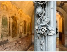 Nahaufnahme eines verzierten Türrahmens mit aufwendigen Rosendekorationen aus Metall; im Hintergrund ein historischer Innenraum mit Deckengewölbe, Wänden mit verblassten Fresken in warmen Farben und einem Marmorsockel.
