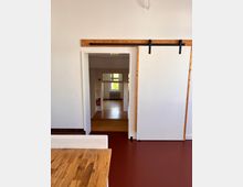 Innenaufnahme eines Raumes mit weiß gestrichenen Wänden, einer hölzernen Schiebetür auf einer Metallführung und einem Blick durch eine Türöffnung in einen angrenzenden Raum mit Holzboden und einem Heizkörper unter einem Fenster. Im Vordergrund ist ein Holztisch auf einem dunkelroten Boden zu sehen.