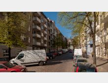 Pflasterstraße in einem Wohngebiet mit mehrstöckigen, teils verputzten, teils verklinkerten Gebäuden und Balkonen links und rechts der Straße. Parkende Autos und Kleintransporter säumen die Straße, während Bäume entlang des Gehwegs frisches Laub tragen.