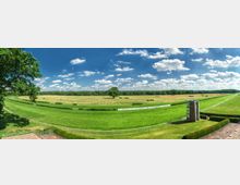 Weitläufige grüne Rennbahn mit gepflegtem Rasen, umgeben von Hecken und einzelnen Bäumen; im Hintergrund ein Waldstreifen und ein blauer Himmel mit vereinzelten Wolken. Im Vordergrund ein Turm und eine Absperrung entlang der Strecke.
