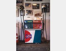 Vintage-Tankstellenautomat mit der Aufschrift „Super Benzin“, umgeben von alten Fotos und Motorsport-Postern an einer Wand. Zwei Zapfschläuche mit Retro-Motorradhelmen sind an den Seiten befestigt, und eine Holzbesen sowie ein Verkehrskegel stehen daneben.